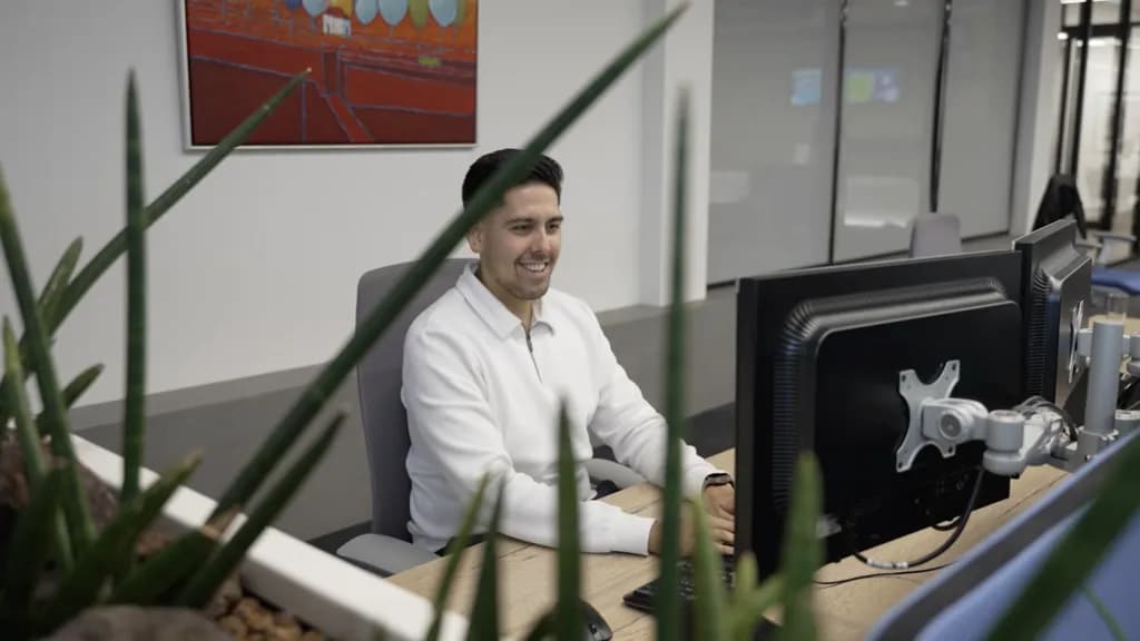 Medewerker van Hoek & Blok werkt lachend achter een monitor op kantoor, gefotografeerd door de planten heen