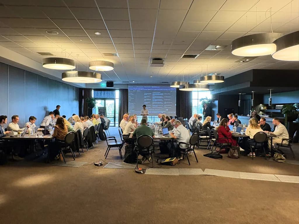 Grote groep professionals volgt een AI-training aan ronde tafels in een evenementenruimte, als beeld bij de Amega case